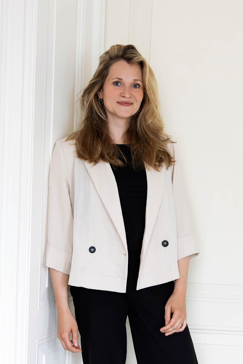 Een persoon met halflang haar draagt een lichte blazer over een zwarte outfit en zwarte schoenen terwijl die tegen een witte muur leunt in een lichte kamer. Beschrijving gegenereerd door AI