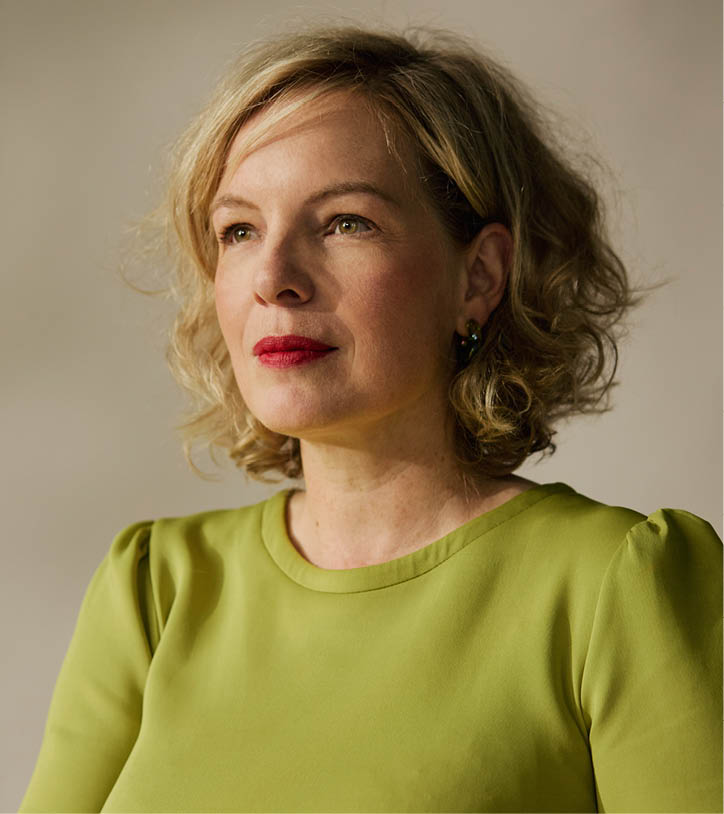 Een persoon met krullend haar draagt een groene blouse en staat voor een neutrale achtergrond. Beschrijving gegenereerd door AI