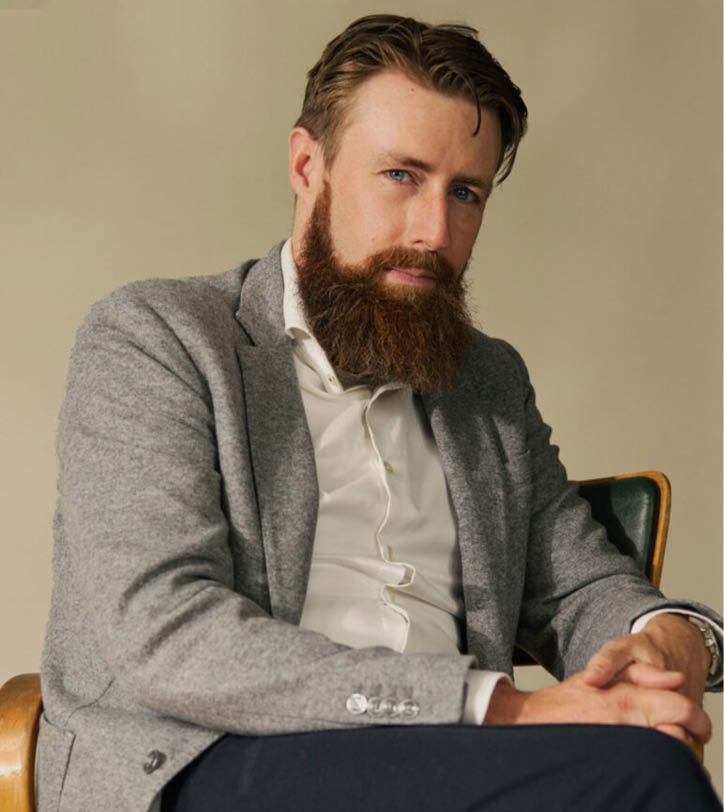 Een persoon met een baard draagt een grijs colbert en een witte blouse en zit met gevouwen handen op een stoel tegen een effen beige achtergrond. Beschrijving gegenereerd door AI