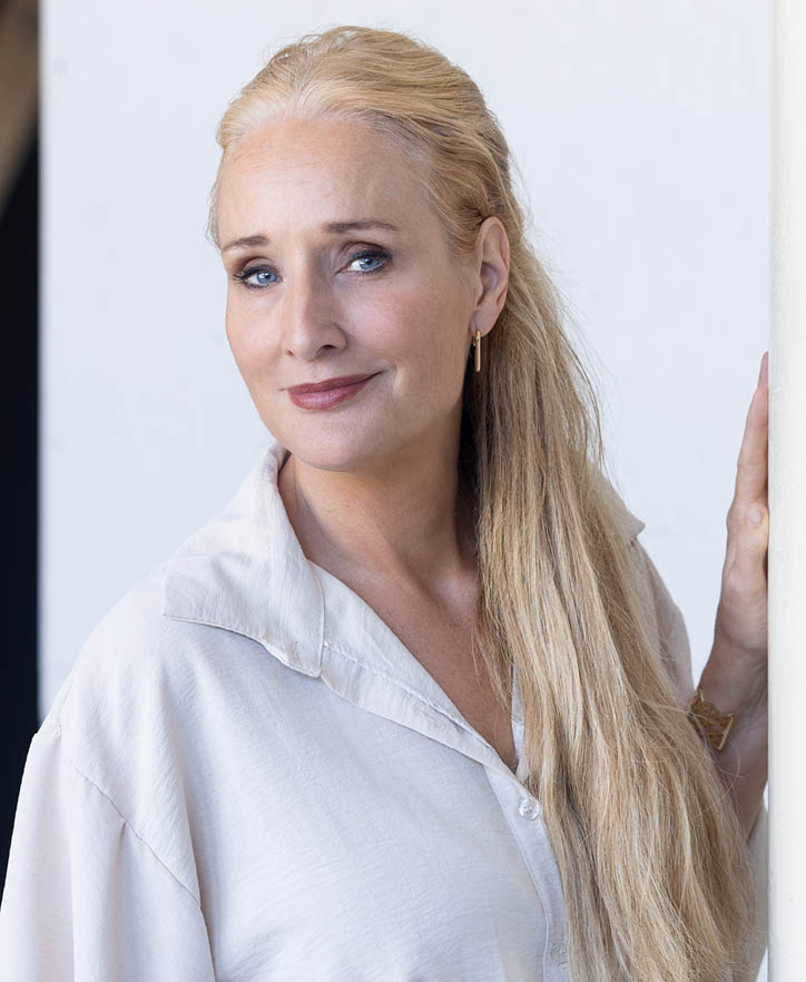 Een persoon met lang, licht haar draagt een lichtgekleurde blouse en een oorbel en glimlacht zachtjes. Beschrijving gegenereerd door AI